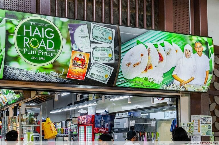 Storefront with a vibrant sign for "Haig Road Putu Piring," since 1985, featuring steamed rice cakes and smiling sellers. Busy interior with customers.