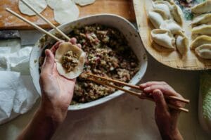 Hands are skillfully using chopsticks to fill a small dumpling wrapper with a savory mixture, surrounded by formed dumplings and an inviting home-cooked atmosphere.