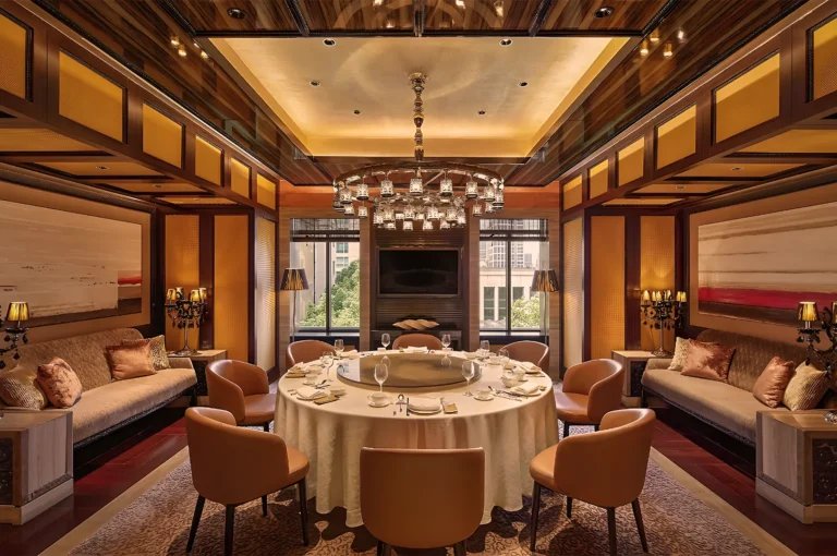 Elegant dining room with a round table set for a meal, plush chairs, and warm lighting. The ambiance is luxurious with modern art and a chandelier.