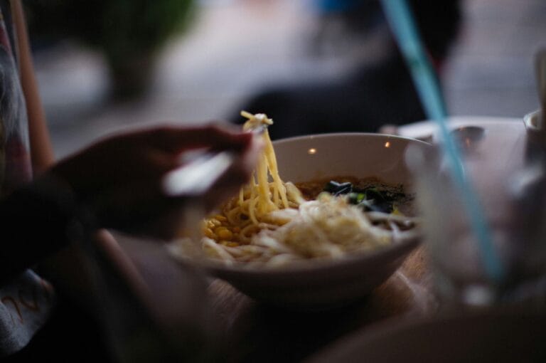 A person with chopsticks lifts noodles from a bowl of ramen. The setting is dim and cozy, conveying a warm atmosphere.
