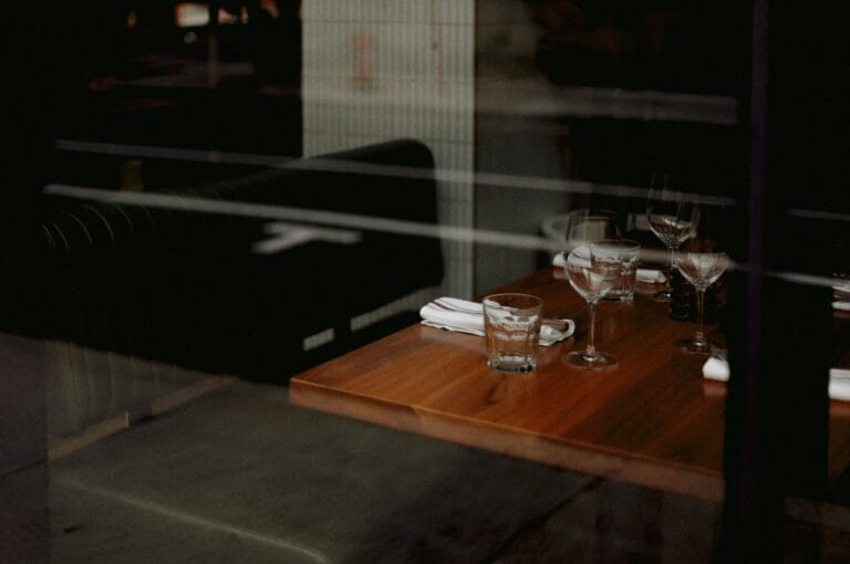 A dimly lit restaurant table set for two with empty glasses, white napkins, and silverware. Cozy ambiance, wooden table, dark cushioned seats.