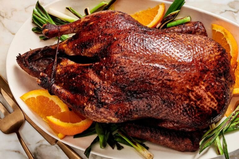 Roasted duck with a crispy, golden-brown skin on a platter, garnished with orange slices and green onions. The setting conveys a warm, appetizing tone.
