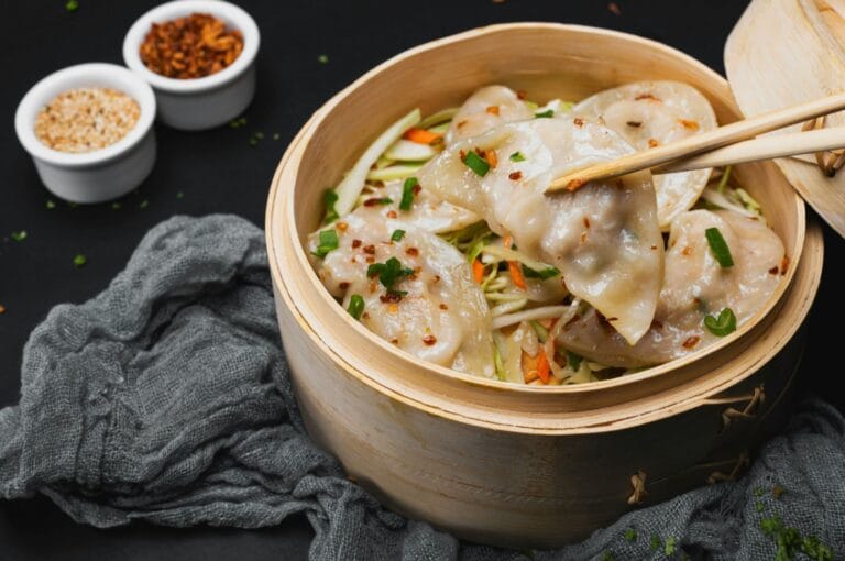 Chopsticks holding a dumpling over a bamboo steamer filled with dumplings and vegetables. Nearby are small bowls of sesame seeds and chili flakes.