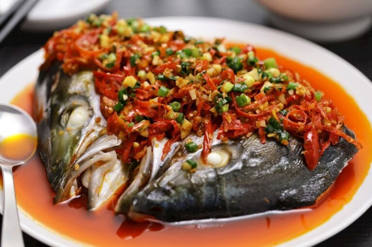 Steamed fish head on a white plate, topped with vibrant red peppers, green onions, and spices, surrounded by a rich, orange sauce. A spoon rests nearby.