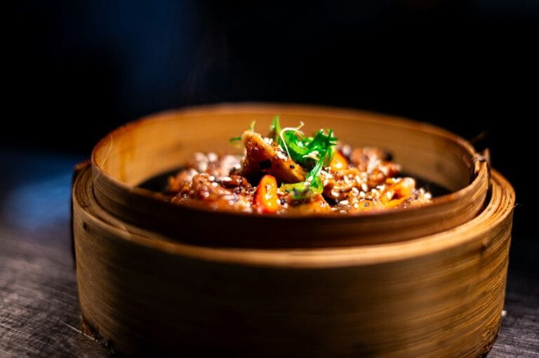 A close-up of a steaming Asian dish in a bamboo basket on a dark background. The dish is garnished with sesame seeds and fresh greens, creating a warm, inviting scene.