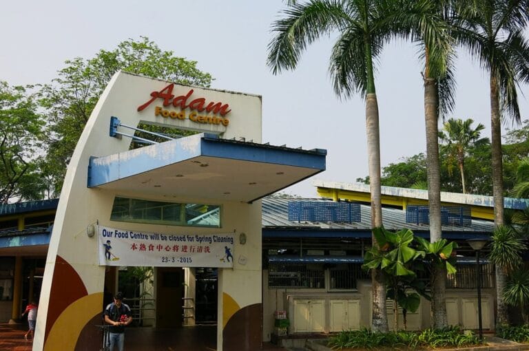 Exterior of Adam Food Centre, featuring a modern entrance with a prominent sign. Surrounded by palm trees, a banner indicates closure for spring cleaning.