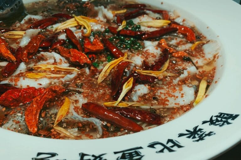 A vibrant dish with red chili peppers, yellow flower petals, and green herbs in broth on a white plate, featuring Chinese characters along the rim. Bold and spicy.