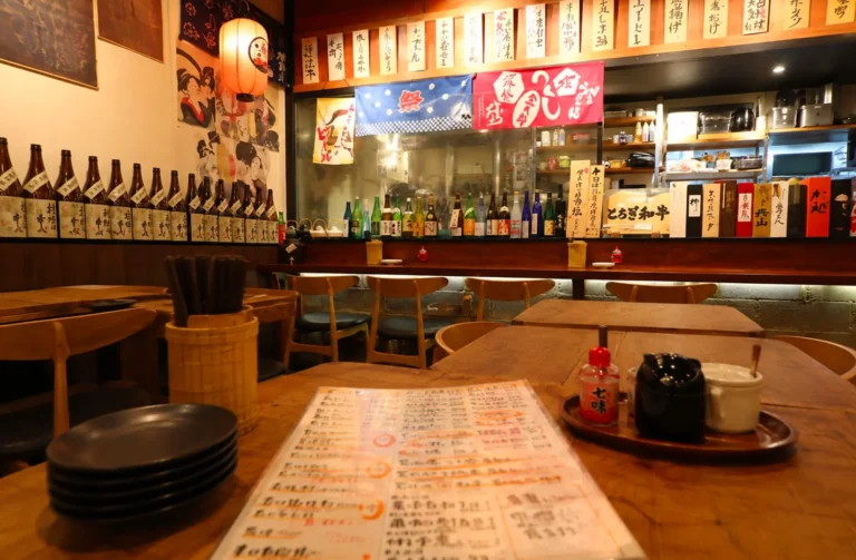 Cozy traditional Japanese izakaya interior featuring a wooden table with a menu, sake bottles lining the bar, and colorful noren curtains