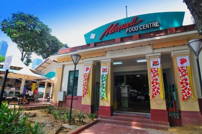 Exterior view of Maxwell Food Centre, a popular market in a city setting. Features include colorful banners, a prominent sign, trees, and outdoor seating.