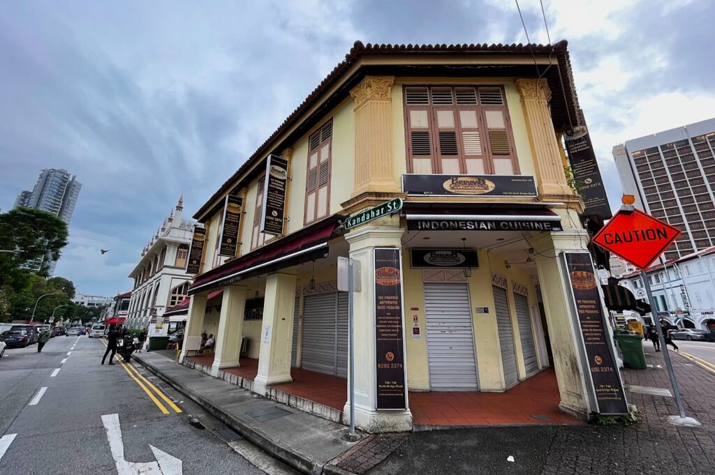 Historic two-story corner building with a sign for Indonesian cuisine. Shuttered doors and a "caution" sign on a cloudy day, creating a quiet, nostalgic feel.