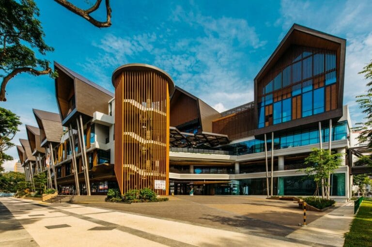 Modern building with a dynamic design, featuring large glass windows, wooden elements, and sloped roofs. Bright blue sky adds vibrancy.