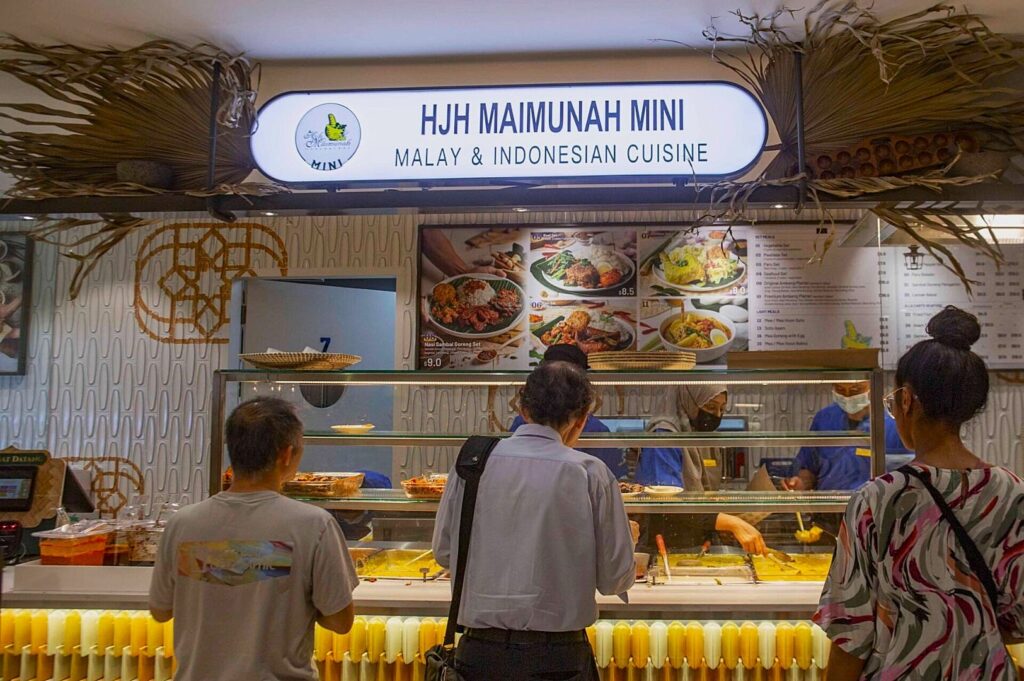 Three people stand at a food stall labeled "HJH Maimunah Mini, Malay & Indonesian Cuisine." The stall displays vibrant dishes, evoking a bustling, appetizing atmosphere.