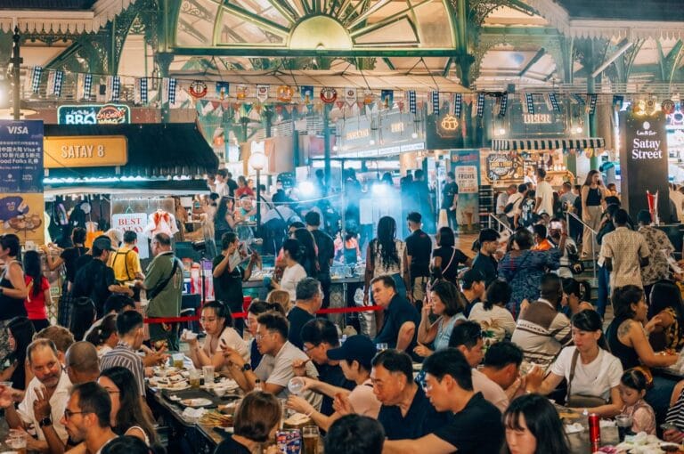 Bustling food market scene with diverse people enjoying meals at crowded tables. Bright lights, food stalls, and festive banners create a lively atmosphere.