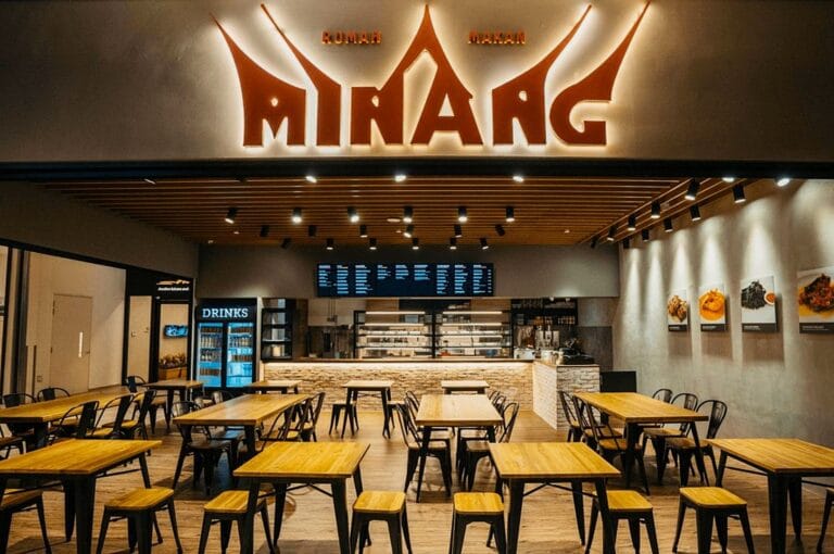 Modern eatery with wooden tables, black chairs, and bright overhead lights. A menu board and food images decorate the back wall, creating a welcoming atmosphere.