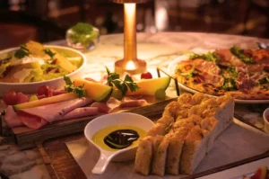 A warmly lit table with a salad bowl, charcuterie board, slices of bread with olive oil and balsamic, and a pizza, creating a cozy dining ambiance.