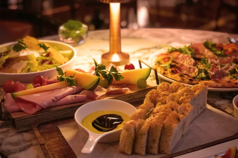 A warmly lit table with a salad bowl, charcuterie board, slices of bread with olive oil and balsamic, and a pizza, creating a cozy dining ambiance.