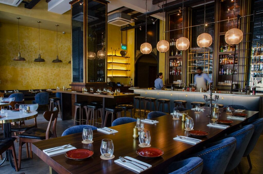 Chic, modern restaurant interior with yellow walls, wooden tables set with glasses and napkins, a well-stocked bar, and soft globe lighting overhead.