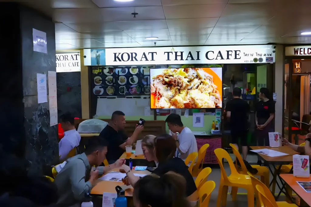 Interior of Korat Thai Cafe with diners seated at tables. A digital menu displays a dish. The atmosphere is casual and bustling.