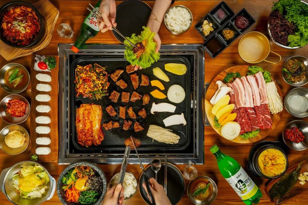 Top view of a Korean BBQ spread with grilling meats, vegetables, and mushrooms. Side dishes, rice, and drinks surround the grill, creating a communal and inviting atmosphere.
