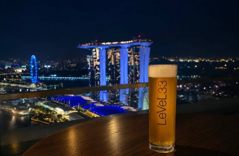 LeVeL33 beer glass on table with nighttime view of Marina Bay Sands and Singapore Flyer illuminated in blue.