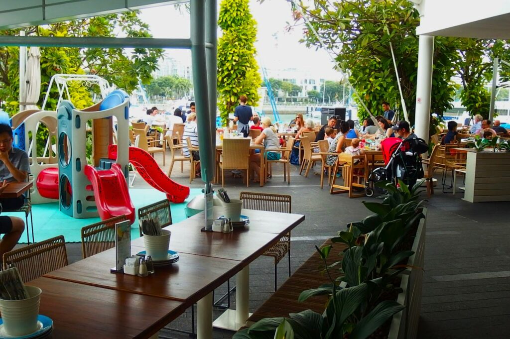 Outdoor restaurant with wooden tables and a lively atmosphere. Diners enjoy meals near a kids' play area with slides. Greenery surrounds the space.