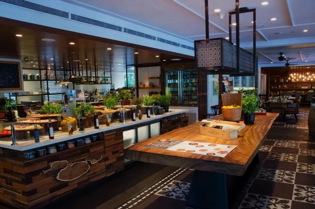 Modern restaurant interior with wooden elements, featuring a long counter adorned with fresh herbs, fruits, and oils. Warm lighting and cozy ambiance enhance the inviting atmosphere.