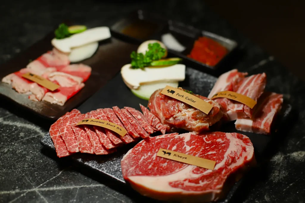 An assortment of raw meats, including ribeye, boneless short ribs, pork collar, and pork belly, displayed on black plates with garnishes. A variety of sauces is visible in the background, creating a gourmet and appetizing presentation.