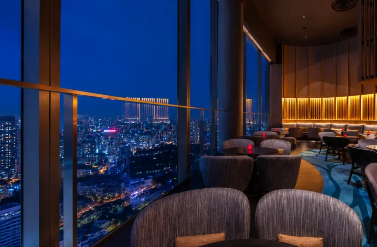 Modern rooftop lounge with plush gray seating and floor-to-ceiling windows overlooking city skyline at dusk with warm golden lighting