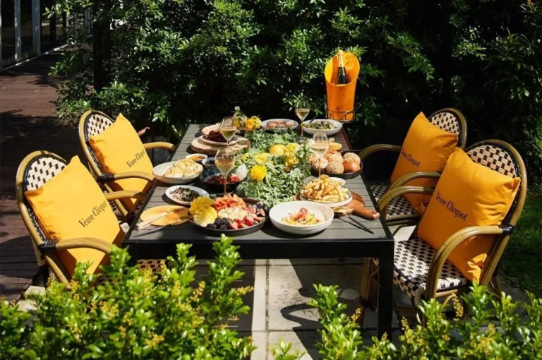 A luxurious outdoor brunch spread on a patio featuring Veuve Clicquot champagne, yellow branded pillows, fresh fruit platters, pastries, and French fries under bright sunlight.