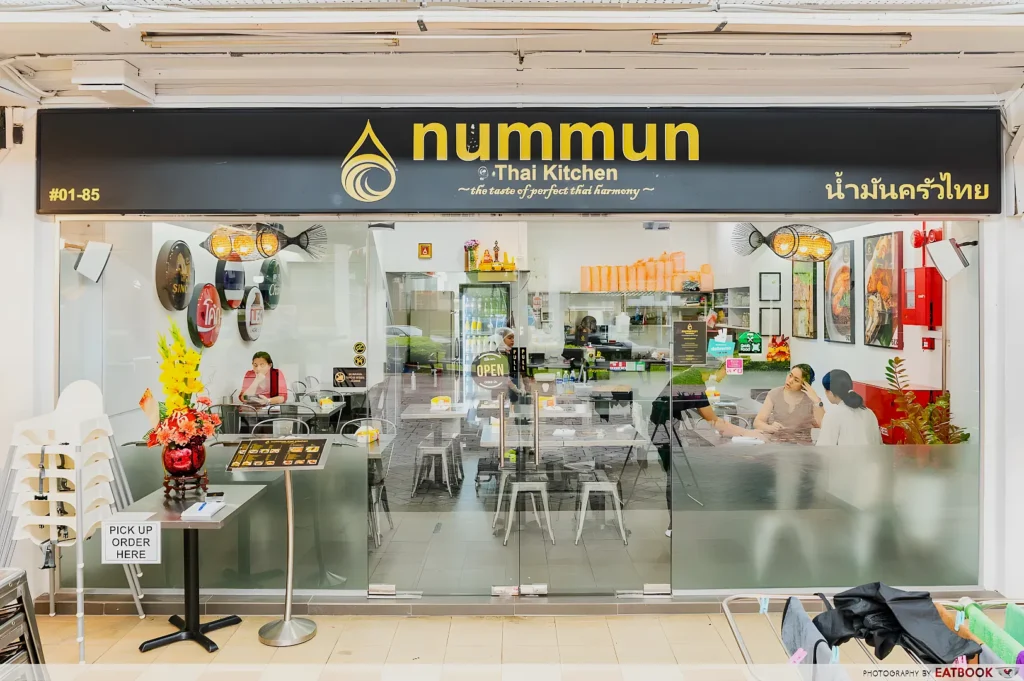 Front view of Nummun Thai Kitchen with glass doors, modern decor, vibrant wall art, and tables inside. Sign reads "Pick Up Order Here." Bright and inviting.