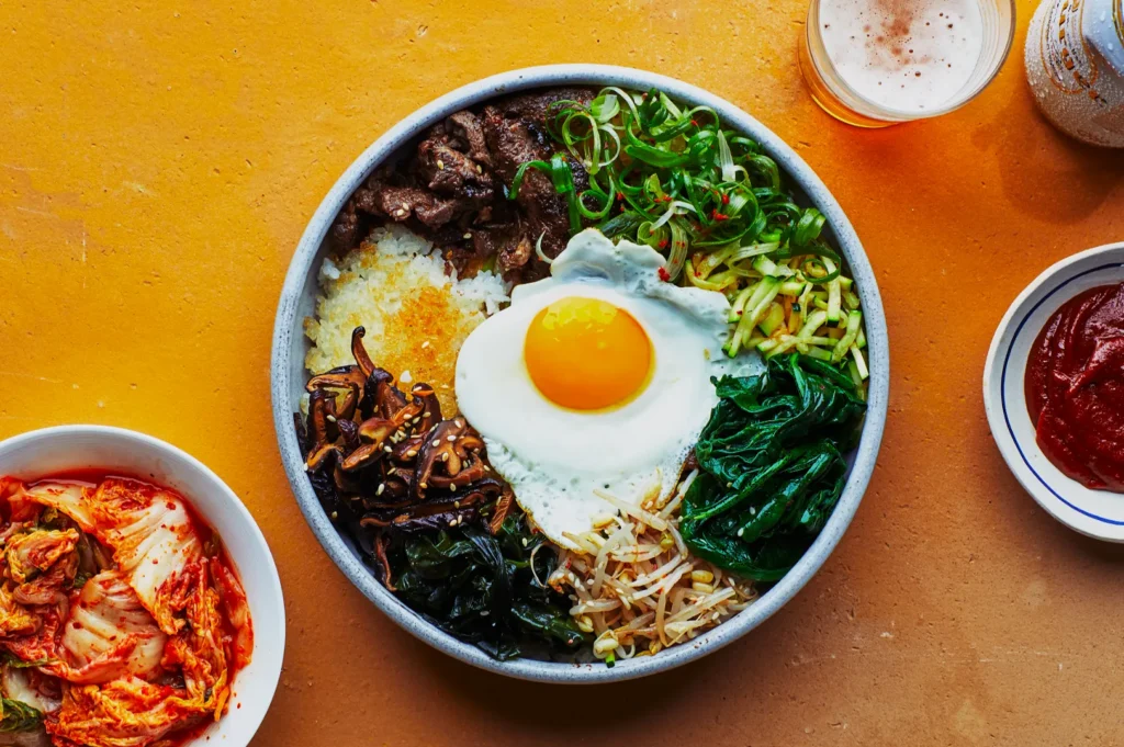 A vibrant bowl of bibimbap with a sunny-side-up egg, beef, and vegetables on rice. Sides include kimchi, red sauce, and beer on a warm yellow table.