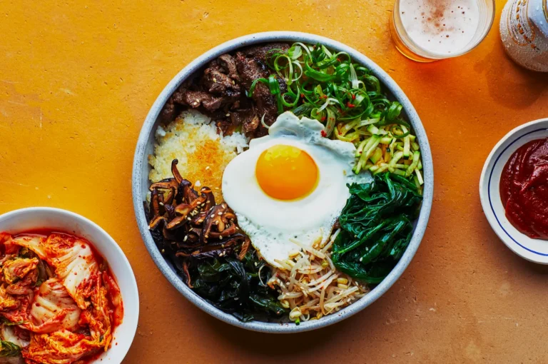A vibrant bowl of bibimbap with a sunny-side-up egg, beef, and vegetables on rice. Sides include kimchi, red sauce, and beer on a warm yellow table.