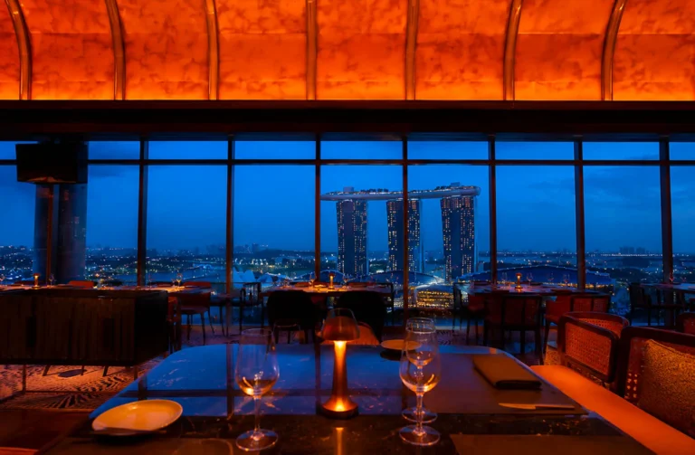 Fine dining restaurant with floor-to-ceiling windows, orange ambient lighting, and tables set overlooking Marina Bay Sands at twilight.