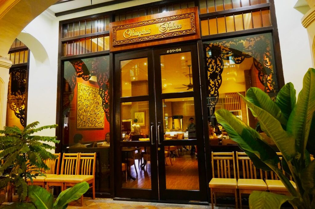 Exterior of a restaurant with a warm, inviting ambiance. Large glass doors reveal a cozy interior with wooden furniture. Ornate signage and lush plants enhance the welcoming feel.
