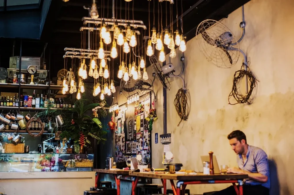Industrial-style cafe with hanging light bulbs, a man working on a laptop at a wooden table. Shelves with bottles and plants in the background. Cozy and creative atmosphere.