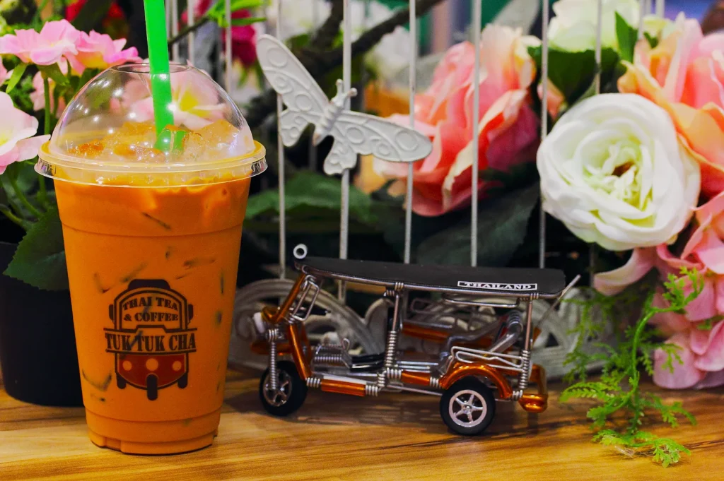 A cup of iced Thai tea with a green straw, labeled "Thai Tea & Coffee Tuk Tuk Cha," sits beside a small tuk-tuk model and pink flowers.