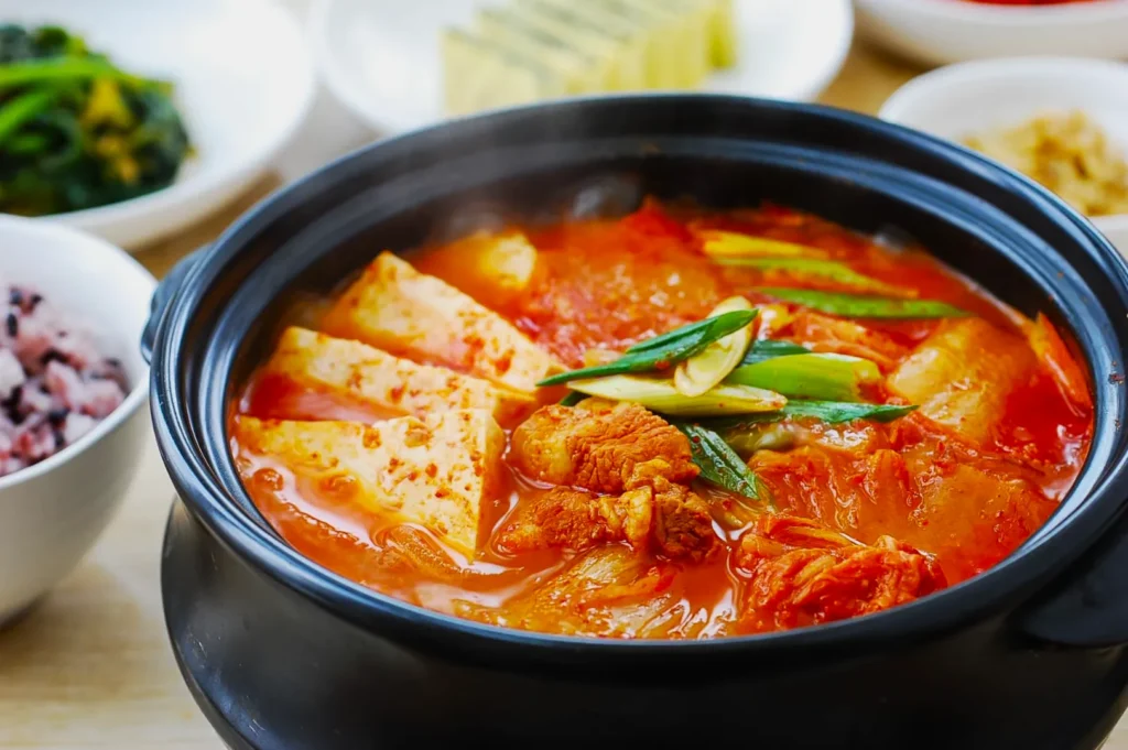 A steaming bowl of spicy kimchi stew with tofu and green onions in a black pot. It’s surrounded by small dishes of rice and side vegetables. Cozy, hearty meal.