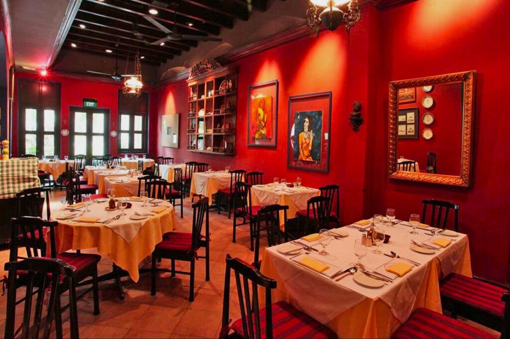 Cozy restaurant with red walls and warm lighting. Tables are set with white linens. Art and a mirror hang on the walls, creating a welcoming ambiance.