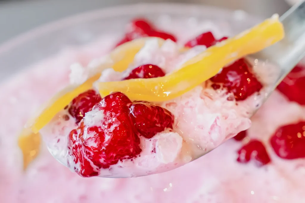 Close-up of a dessert spoon with red strawberries, yellow mango strips, and shaved ice in creamy pink sauce, conveying a sweet and refreshing treat.