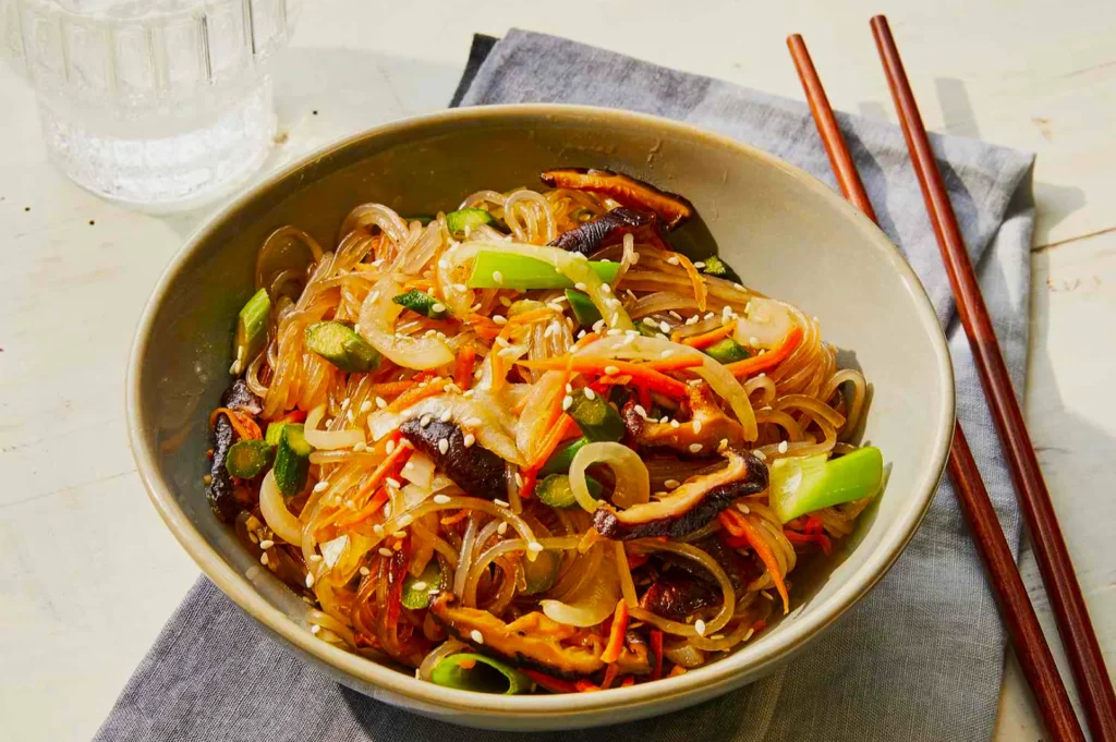 A bowl of colorful Korean japchae with glass noodles, vibrant vegetables, and mushrooms, garnished with sesame seeds. Chopsticks rest beside the bowl.