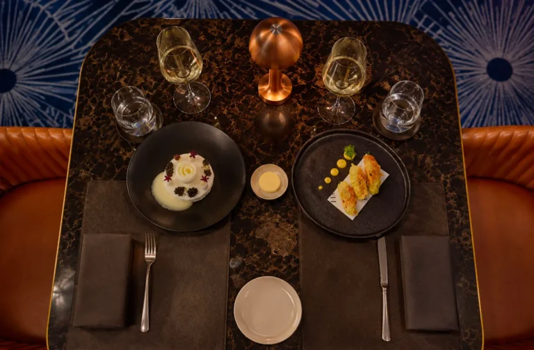 Overhead shot of an elegant dining table for two featuring plated gourmet appetizers and dessert, wine glasses, and a copper lamp on dark marble