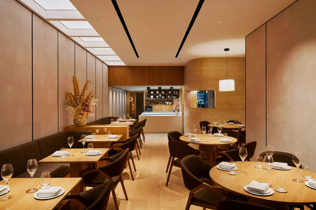 Elegant restaurant interior with wooden tables, set with glasses and white plates. Soft lighting, minimalist decor, and tall pampas grass add a modern, serene ambiance.