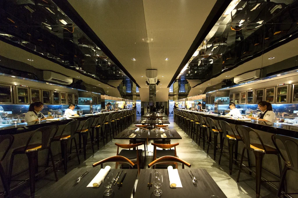 Sleek, modern restaurant interior with a long bar, chefs preparing dishes, wood-accented tables and chairs, and a reflective ceiling enhancing the elegant ambiance.
