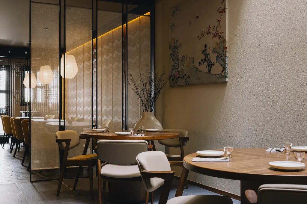Modern restaurant interior with round wooden tables, beige chairs, and decorative glass panels. Warm lighting and a serene, elegant ambiance.