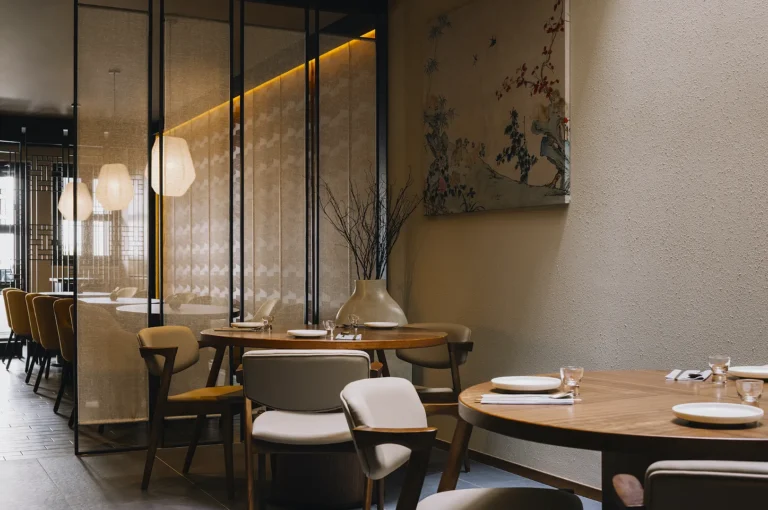 Modern restaurant interior with round wooden tables, beige chairs, and decorative glass panels. Warm lighting and a serene, elegant ambiance.