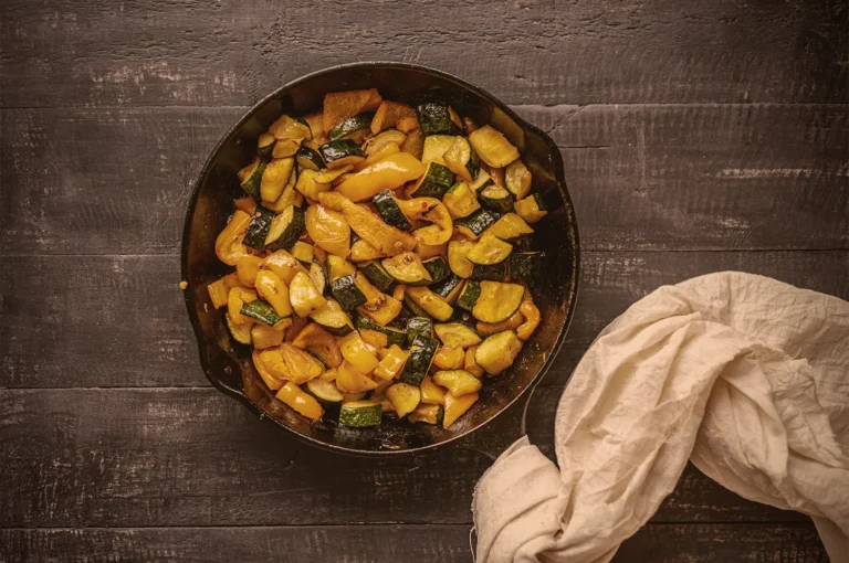 A skillet with sautéed zucchini and yellow bell peppers on a dark wooden surface. A light cloth is casually placed beside the skillet.