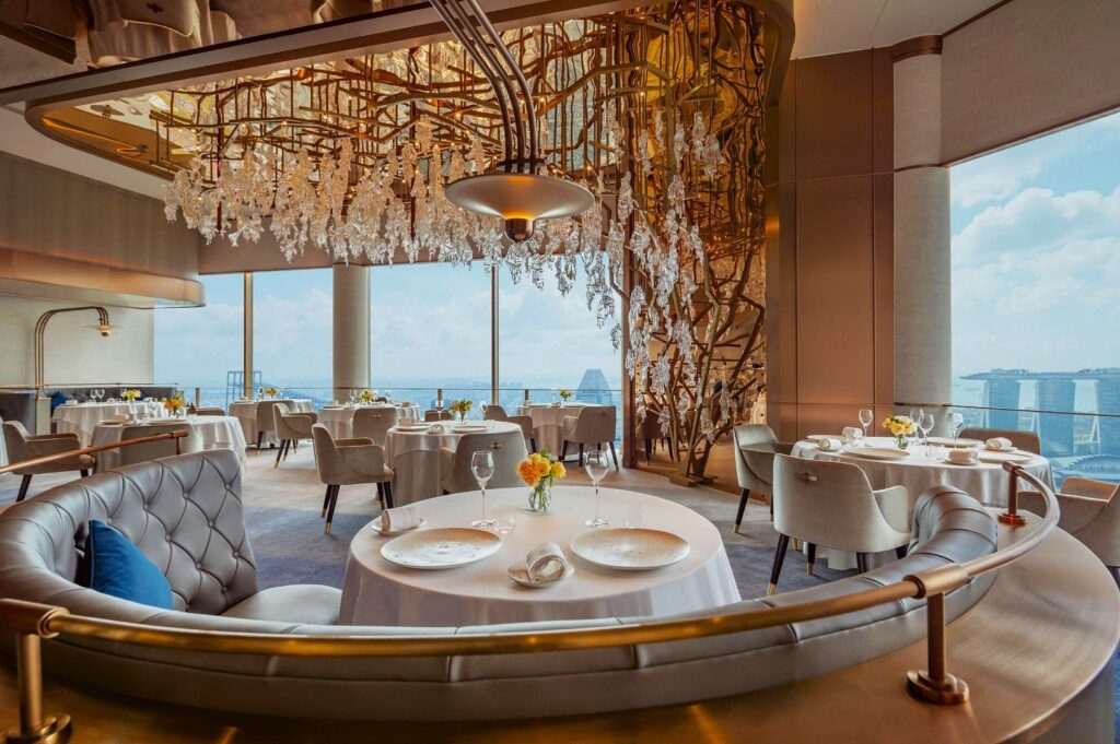 Elegant restaurant interior with round tables set for dining, plush seating, and large windows showcasing a cityscape. Crystal chandelier adds sophistication.