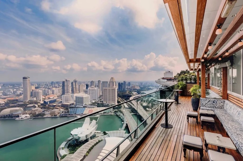 Rooftop terrace with wooden decking overlooking a city skyline, featuring modern skyscrapers and a waterfront. The scene is bright and airy.