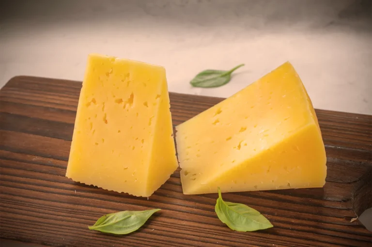 Two wedges of yellow cheese with visible holes, resting on a wooden board. Fresh basil leaves are scattered around, creating a rustic atmosphere.