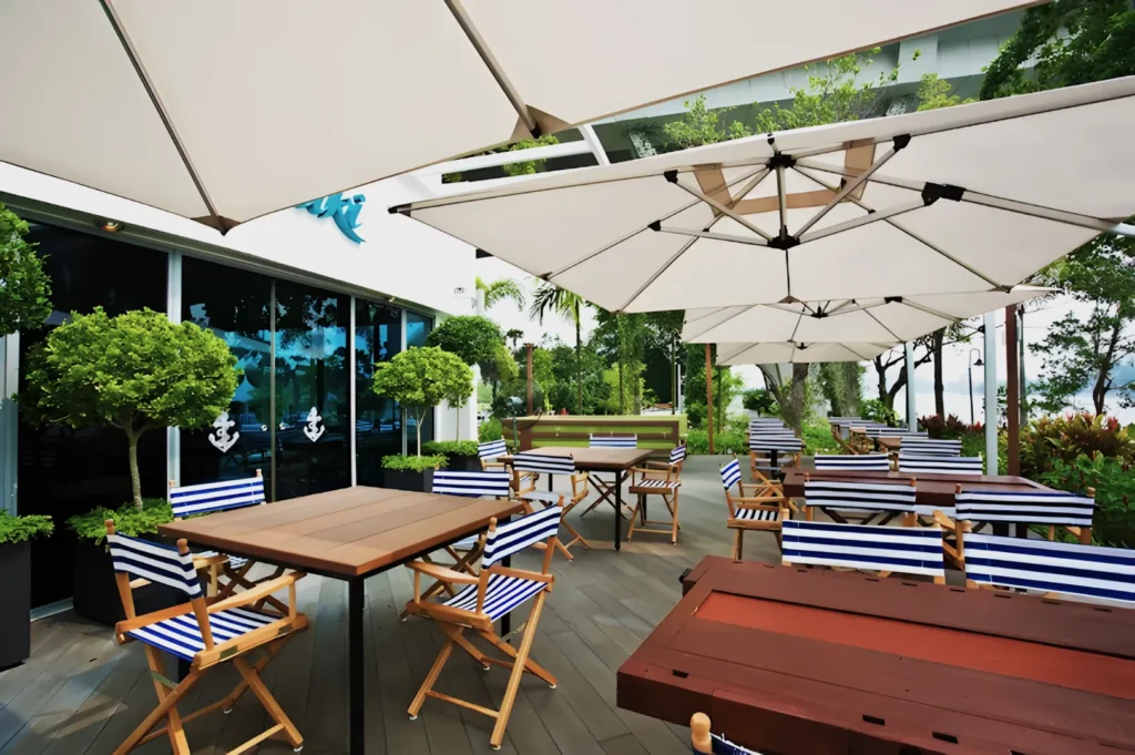 Outdoor café with striped chairs, wooden tables, and large white umbrellas. Surrounded by greenery, the setting feels inviting and serene.
